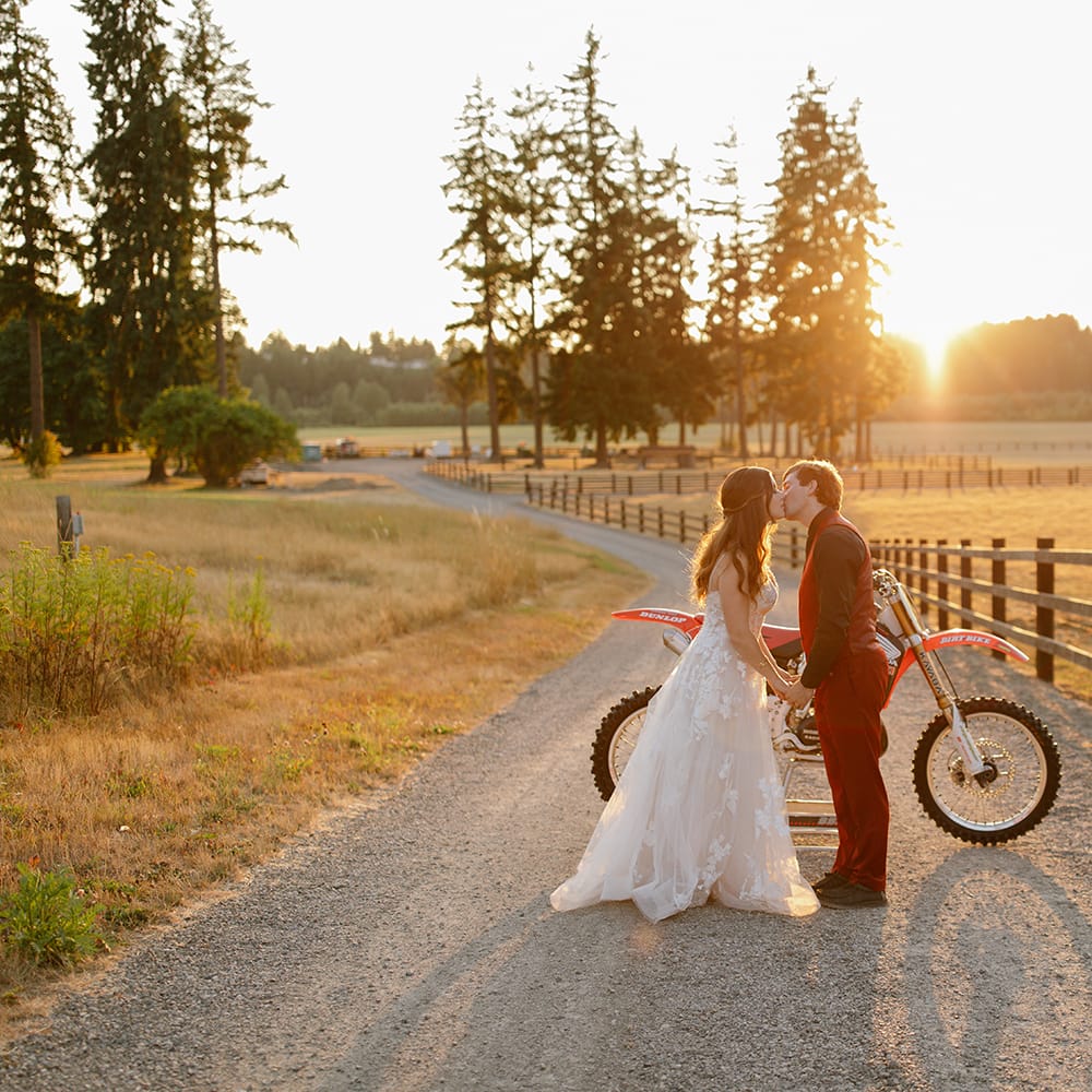Bride in elegant gown and groom in red suit kiss at sunset with dirt bike in background