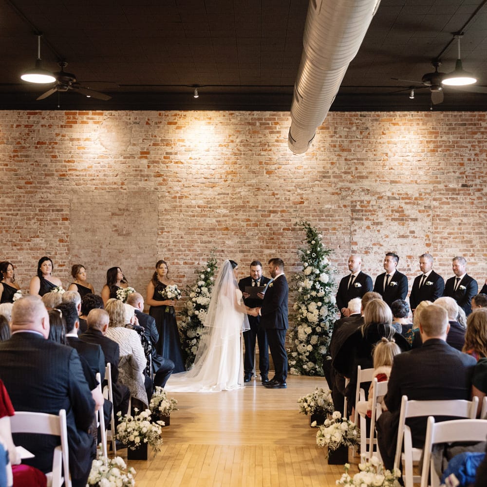 A bride and groom exchange vows in front of a rustic brick wall adorned with asymmetrical white floral arrangements at Liederkranz.