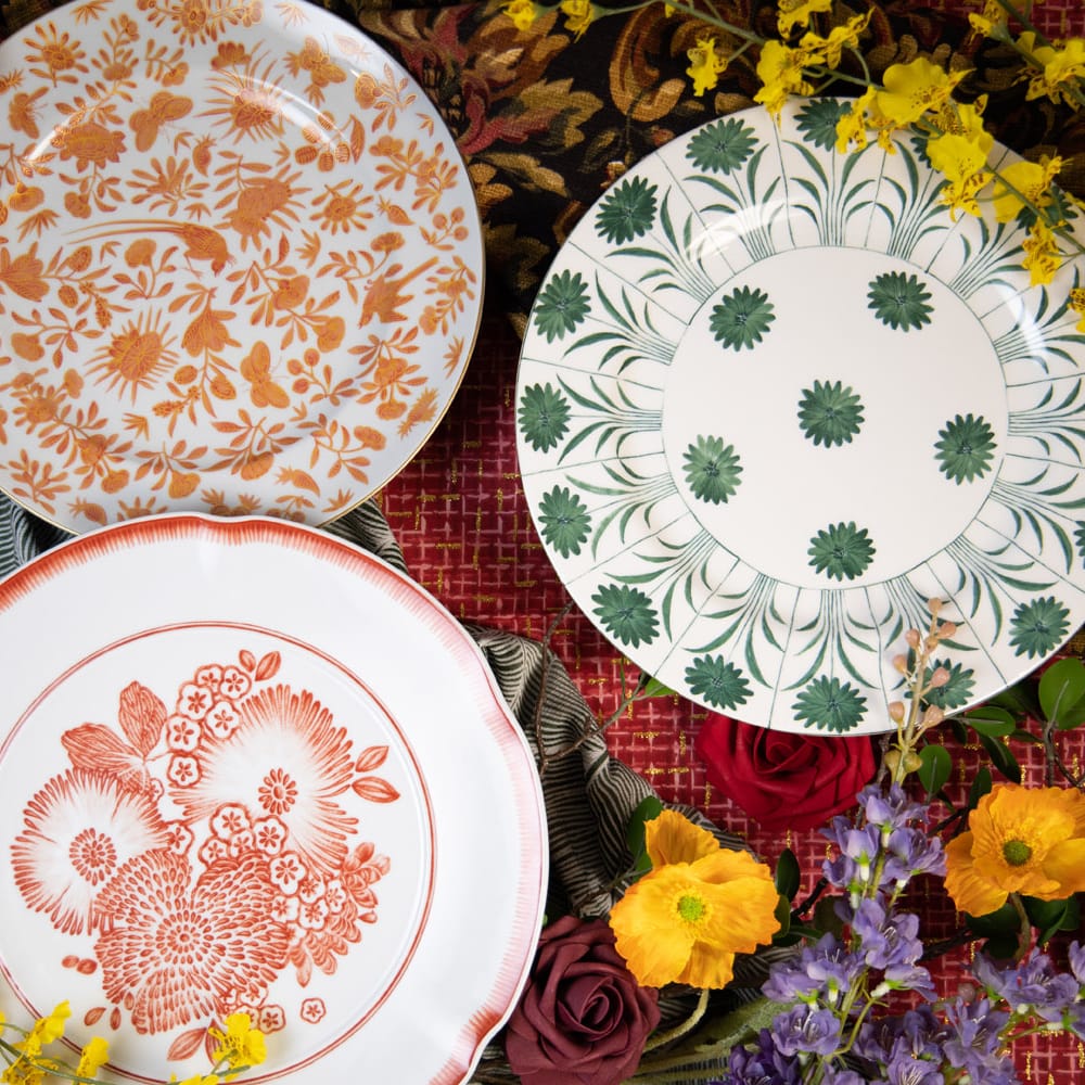 Three designer plates from Maison de Carine—orange Sacred Bird and Butterfly, green Topiary, and coral Oscar’s Coral—styled with roses, poppies, and patterned linens