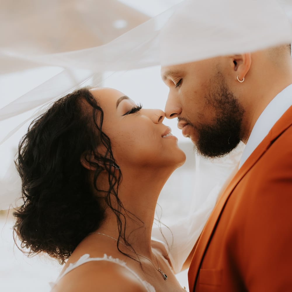 Bride and groom share an intimate moment under the bride’s veil on their fall wedding day, with the groom in a burnt orange suit and the bride in a white lace gown.