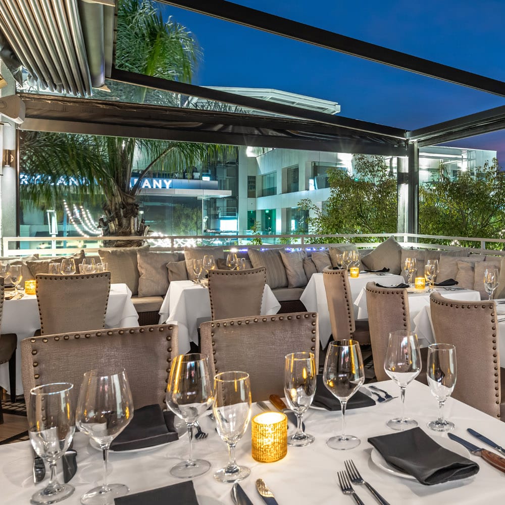 Rooftop dining terrace at Mastro’s Beverly Hills set for an evening event with white linens, candlelight and plush seating under a retractable roof.