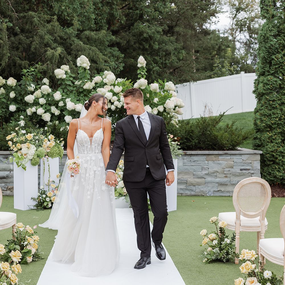 Garden wedding ceremony inspiration featuring a couple walking down a white aisle lined with floral arrangements and rattan chairs during a summer styled shoot at The Hutton House in Minnesota