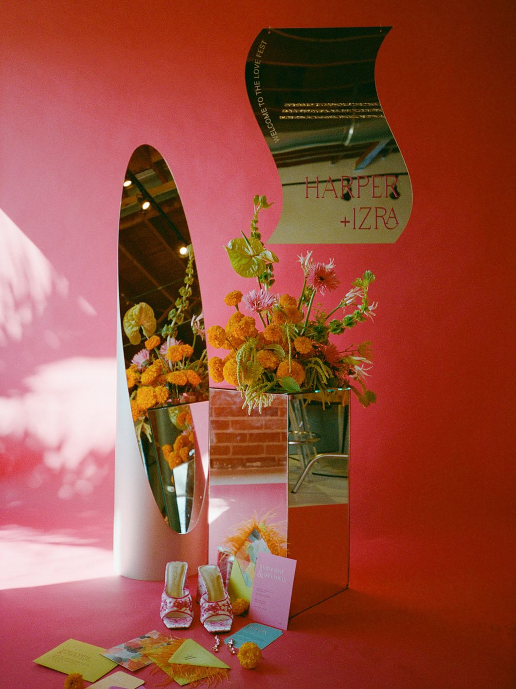 Maximalist wedding vignette with sculptural mirrors, orange florals, colorful stationery and floral bridal shoes against a bold coral backdrop