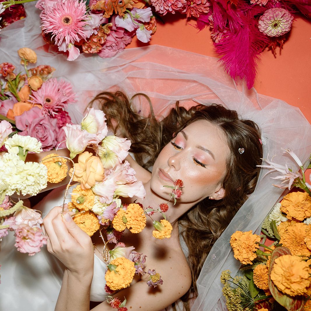 Bride surrounded by vibrant pink and orange wedding flowers while holding a cascading floral arrangement in a playful editorial portrait