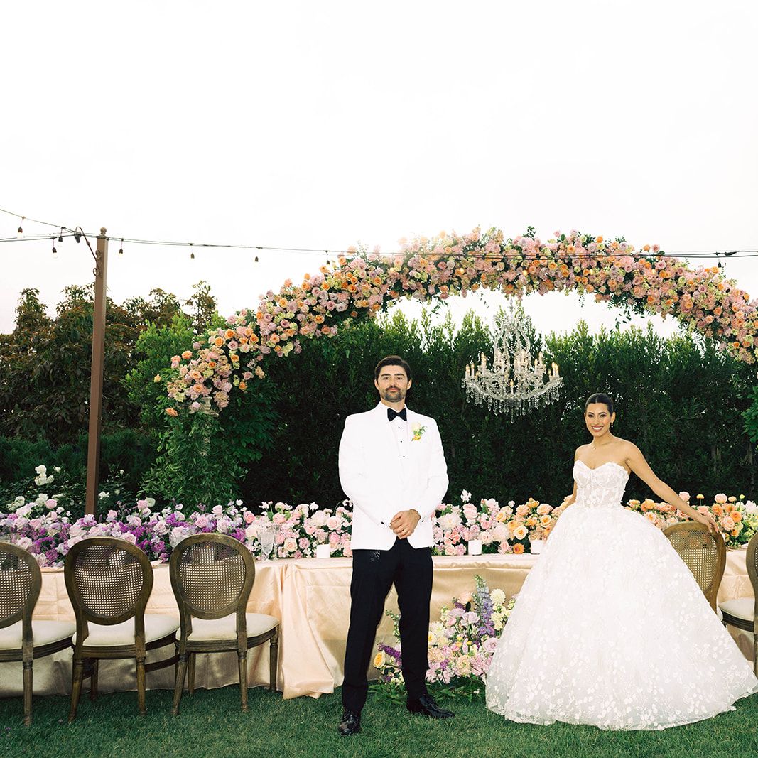 Former Miss California Natasha Alexis Nichelman and Zach Nichelman pose at their reception's head table under a 15-foot floral arch with cascading flowers and a chandelier at Tuscan Rose Ranch.