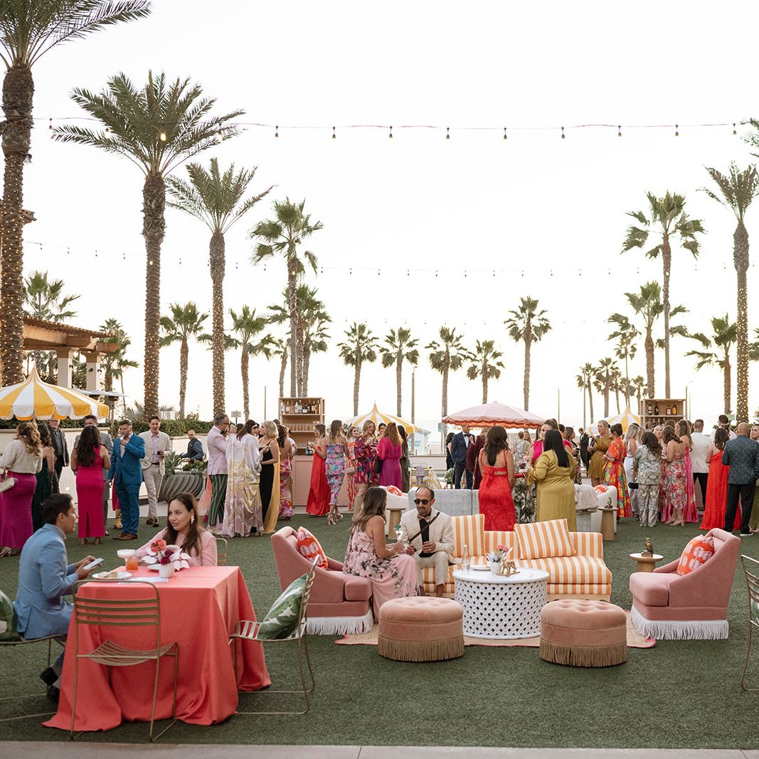 Guests at the Curate Gala 2024 enjoy an elegant outdoor cocktail hour with vibrant lounge furniture, striped umbrellas, and palm tree views at the Waterfront Beach Resort in Huntington Beach