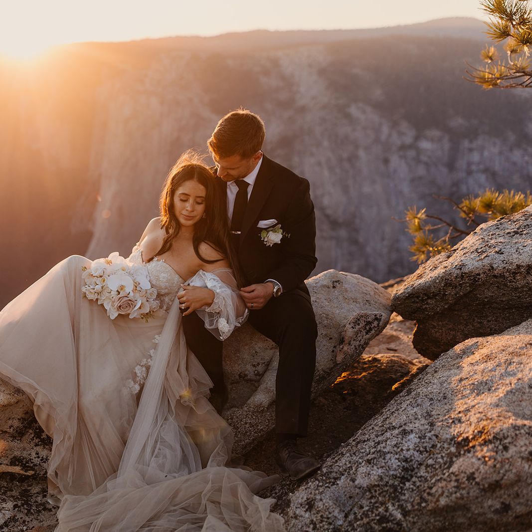 Bride and groom sitting together on a rock at Glacier Point in Yosemite National Park, with the sun setting behind them, creating a golden glow.
