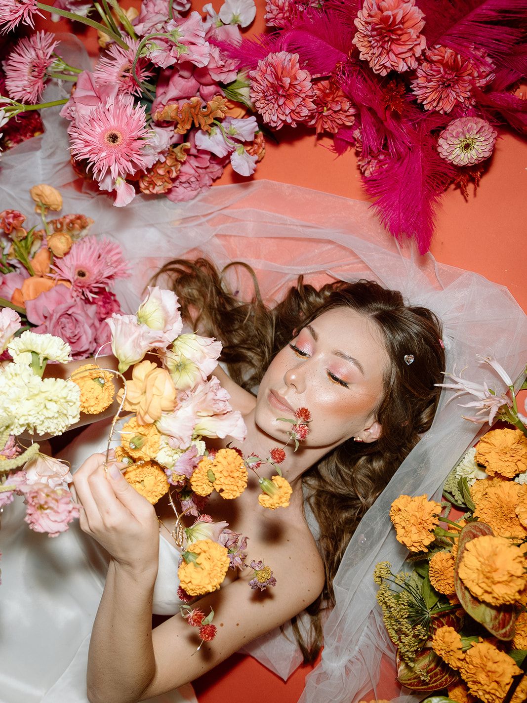 Bride surrounded by vibrant pink and orange wedding flowers while holding a cascading floral arrangement in a playful editorial portrait