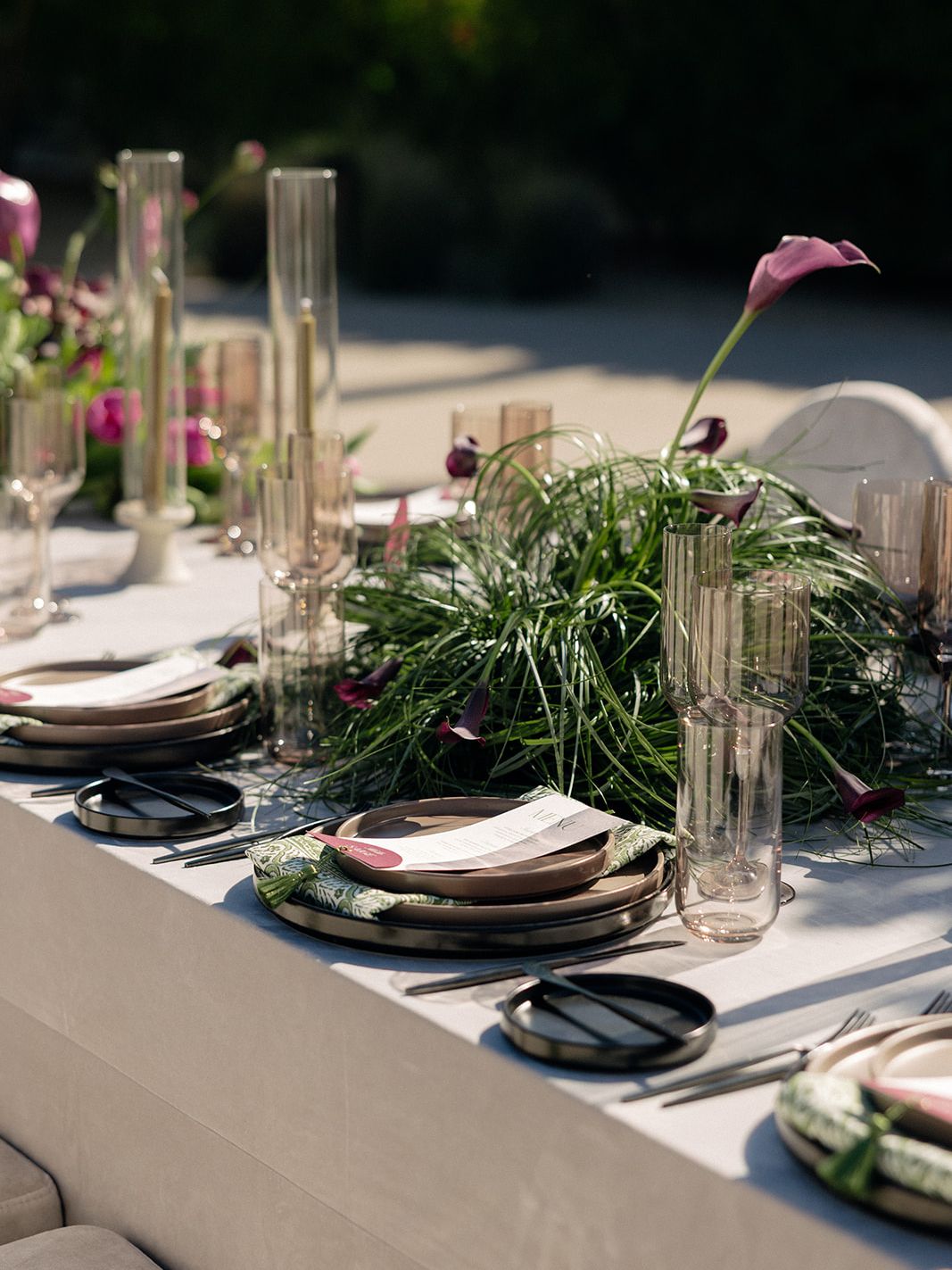 Outdoor wedding tablescape with sculptural grasses, purple calla lilies, neutral linens, modern place settings and tall candles