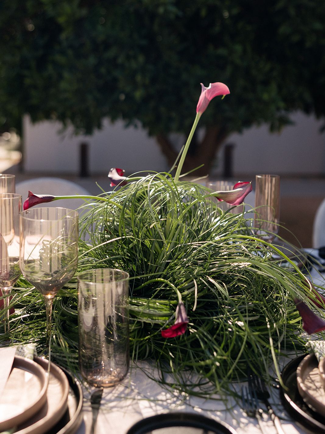 Sculptural wedding centerpiece of flowing green grasses and purple calla lilies surrounded by smoky glassware and modern place setting