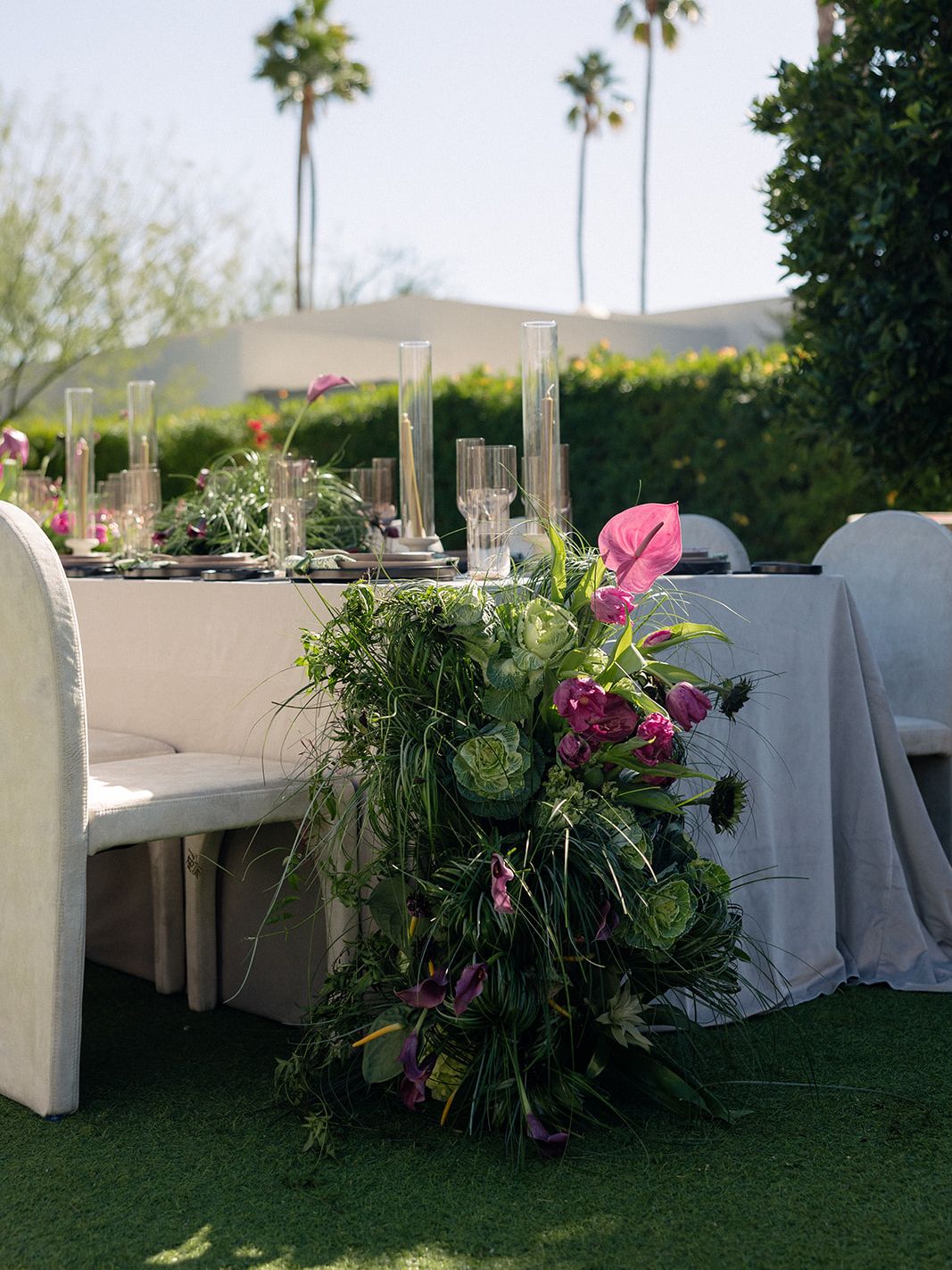 Organic floral installation with ornamental cabbage, pink and purple flowers and cascading grasses beside an outdoor reception table