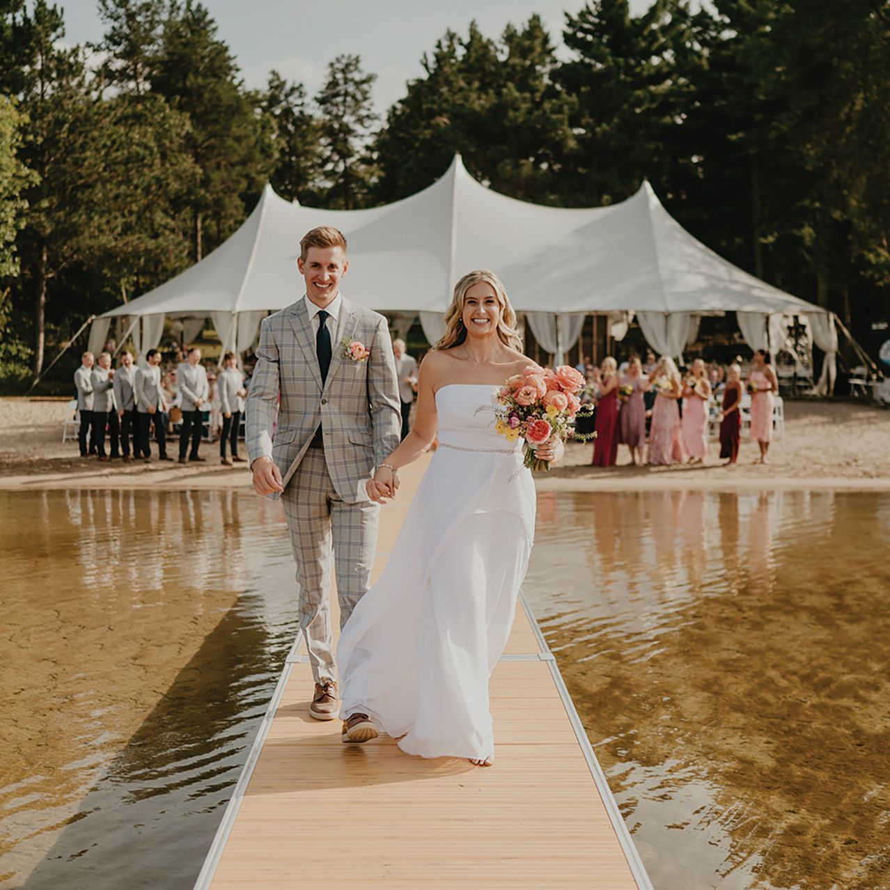 Bride and groom stand holding hands at the end of a dock with wedding party and tented reception behind them in lakeside wedding day