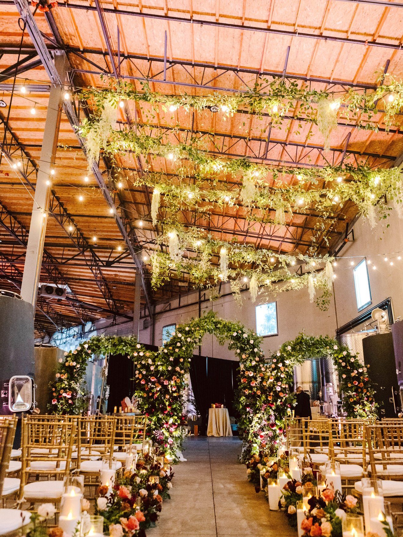 Wedding ceremony aisle lined with flowers and candles, leading to three floral arches beneath greenery and string lights