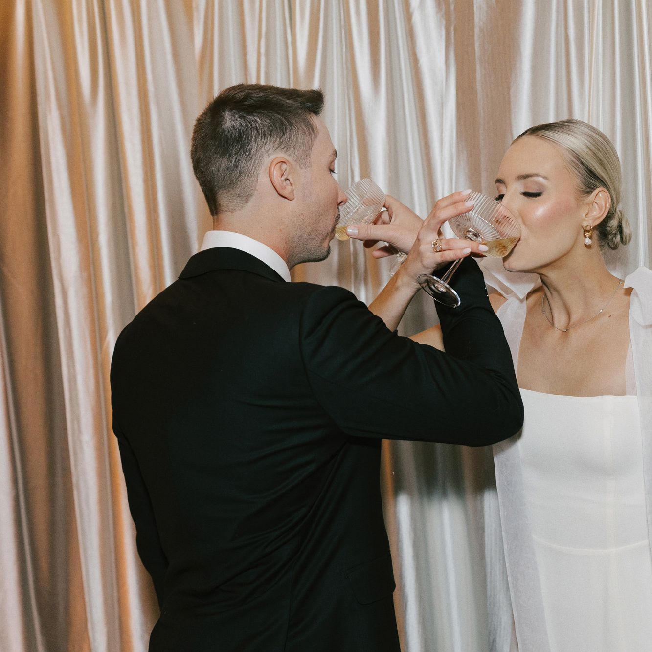 Bride and groom intertwine arms, sipping champagne against a draped backdrop