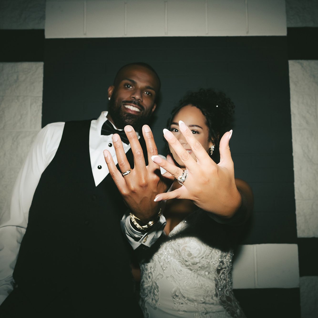 Bride and groom flaunt their rings, holding their hands up as they stand against a black and white checkered background