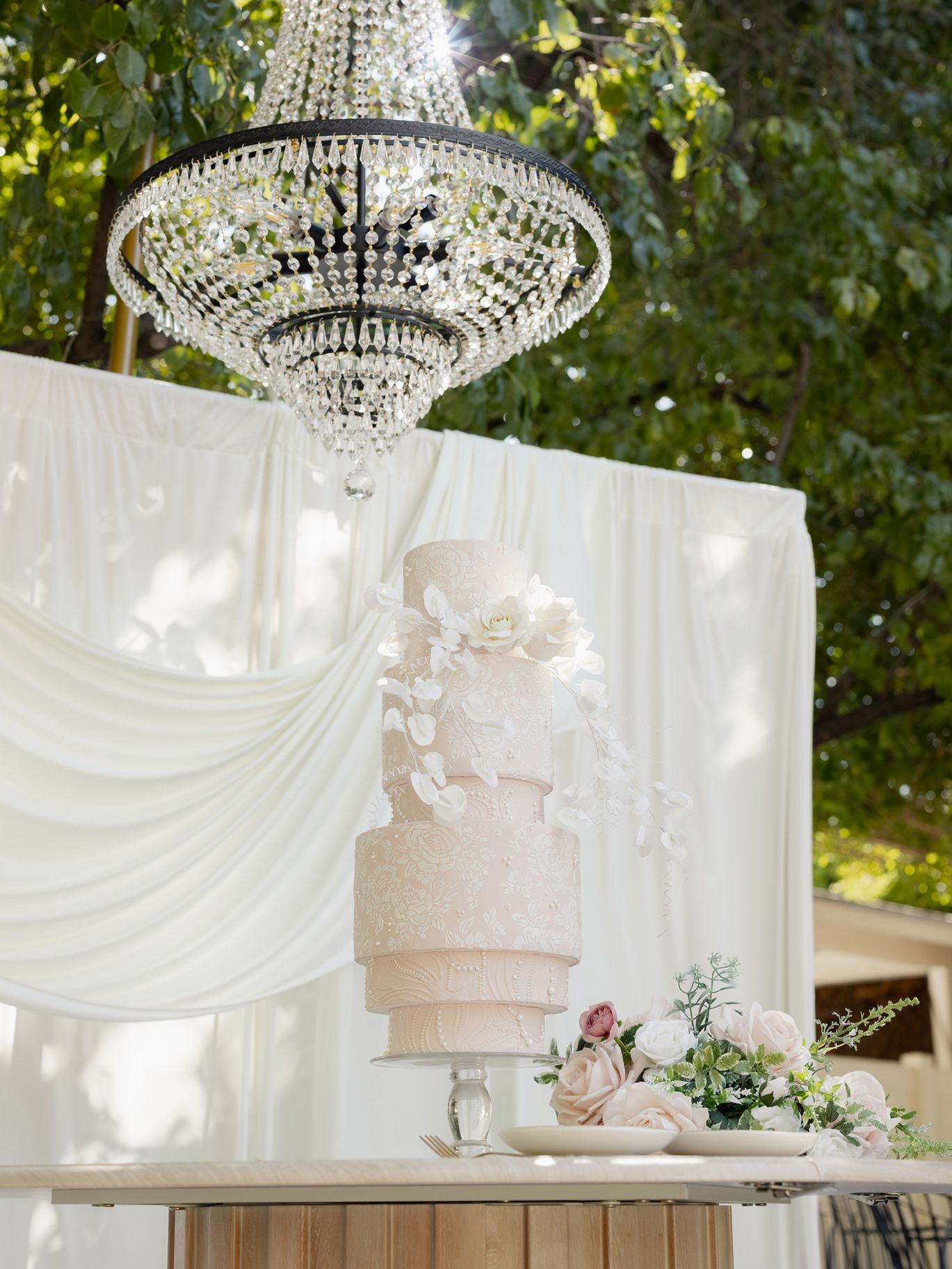Blush pink tiered wedding cake with white floral details beneath an outdoor crystal chandelier
