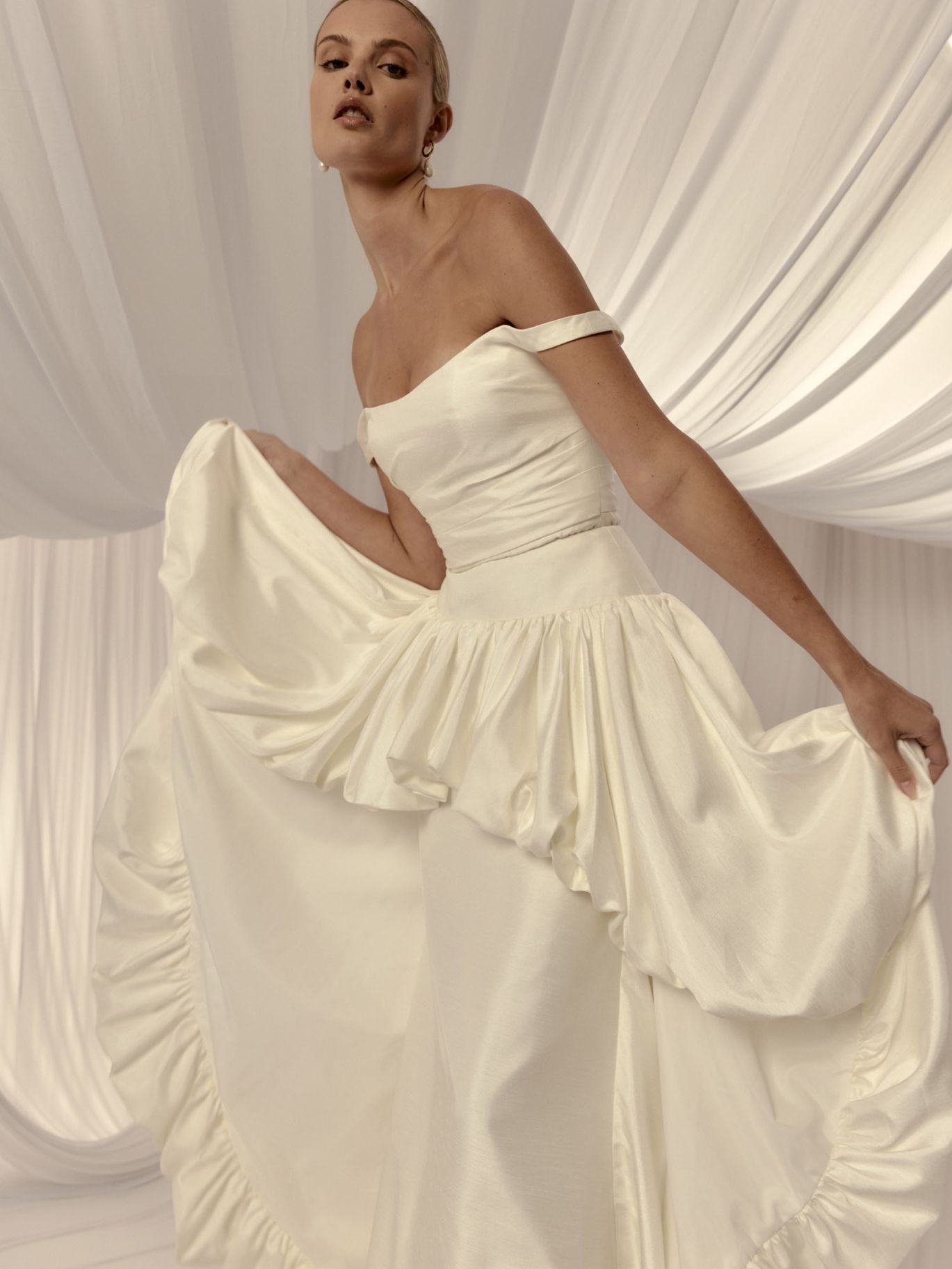 Hera Couture Marguerite wedding dress with a fitted lace gown and off-the-shoulder satin draping, featured in a romantic draping trend roundup from New York Bridal Fashion Week.
