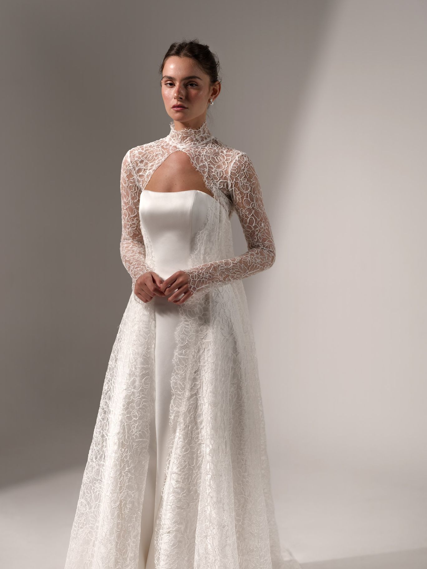 Lihi Hod Muse Topper from the Fall 2027 bridal collection, featuring a high-neck floral lace overlay, long sleeves and a front cutout over a structured wedding dress.