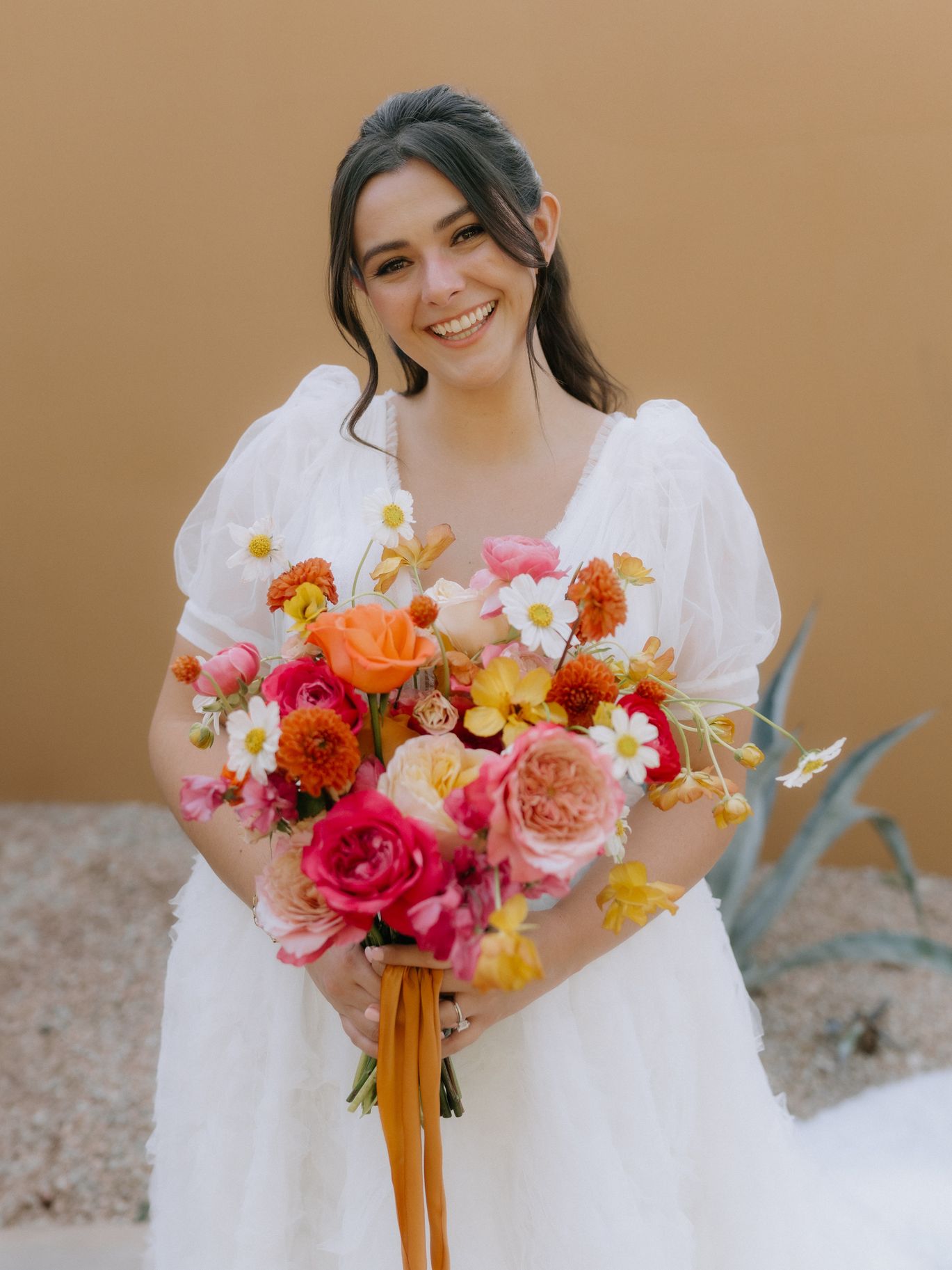 Smiling bride in a white puff-sleeve wedding dress holds a colorful garden-style bouquet of pink, orange and yellow blooms by Fleur de Vie Studio
