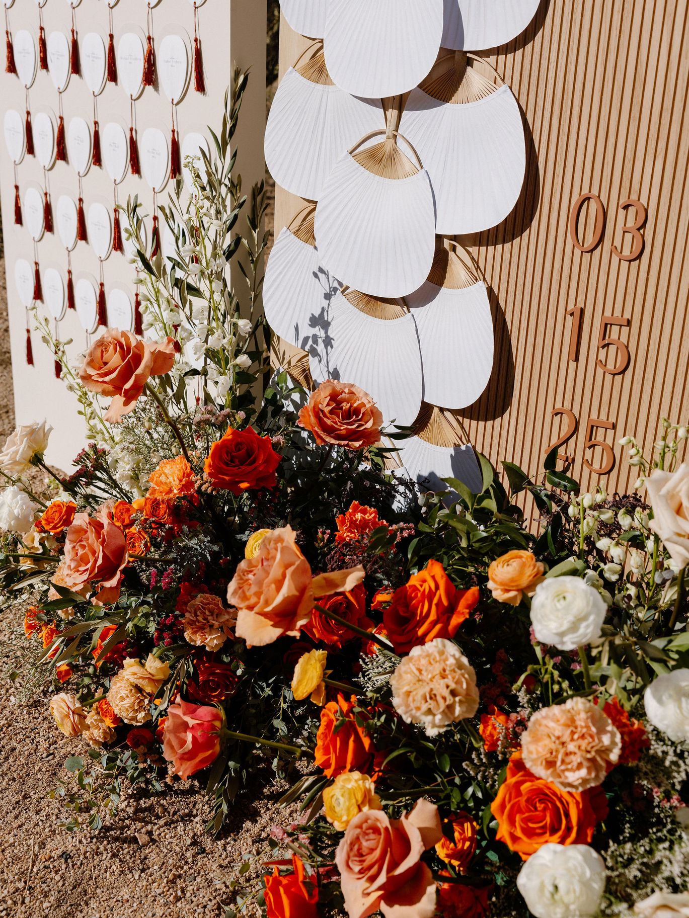 Close-up of orange, peach and white wedding flowers beneath a wood escort card display decorated with white fans