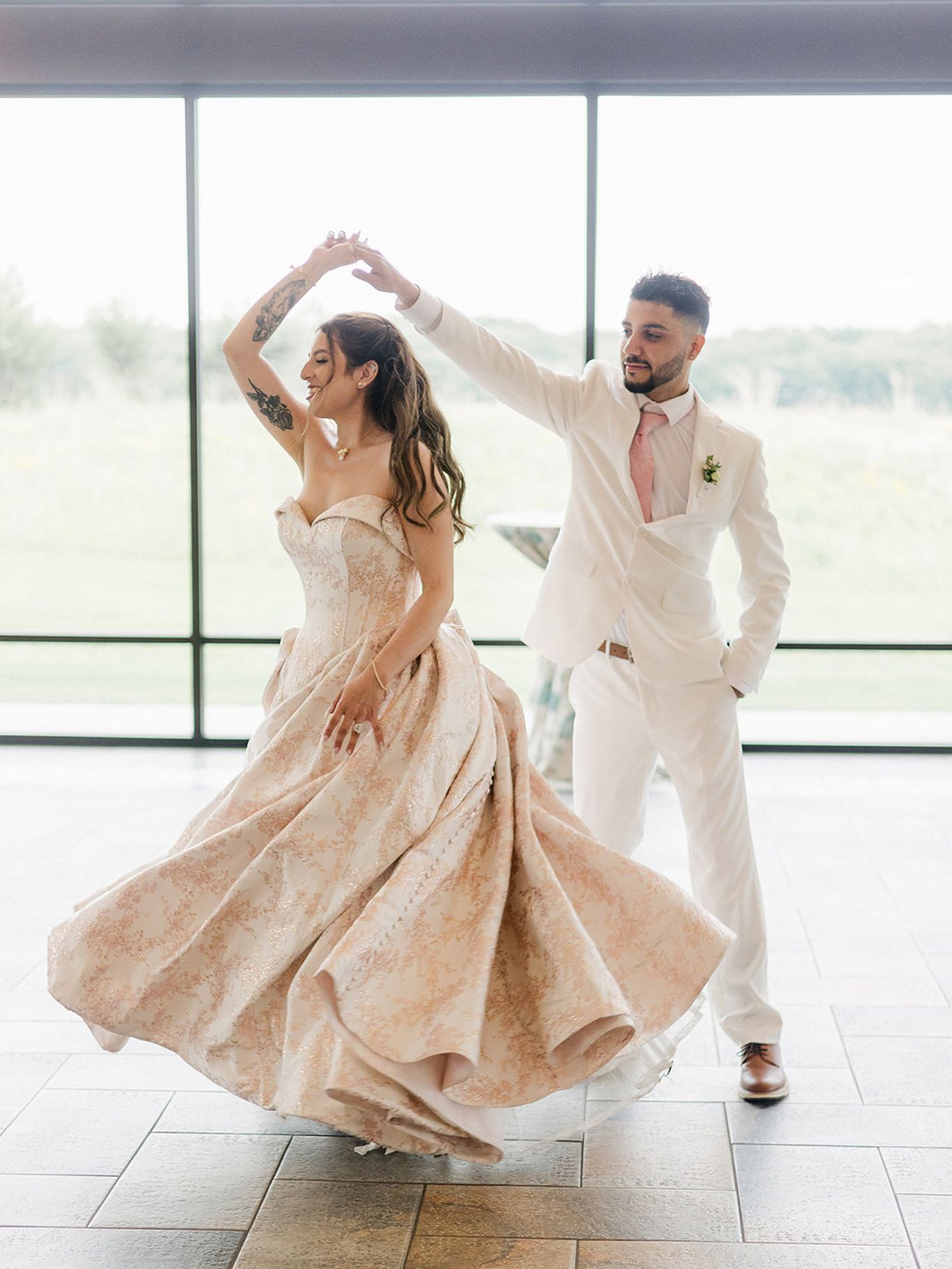 Groom in a white suit twirls his bride in a voluminous blush-patterned wedding gown in front of floor-to-ceiling windows.