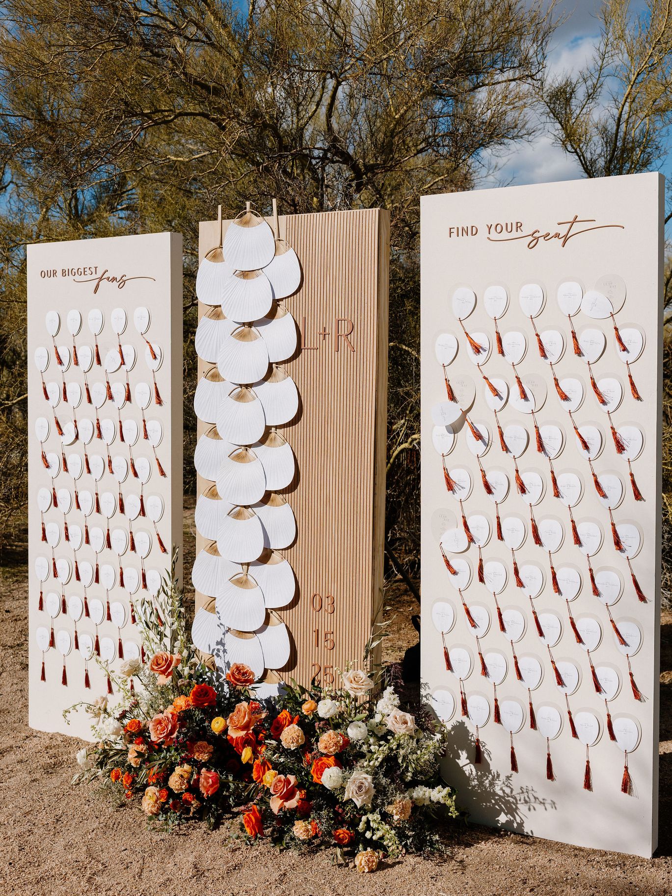 Outdoor wedding escort card display with white fan-shaped place cards, rust tassels and orange, peach and white flowers