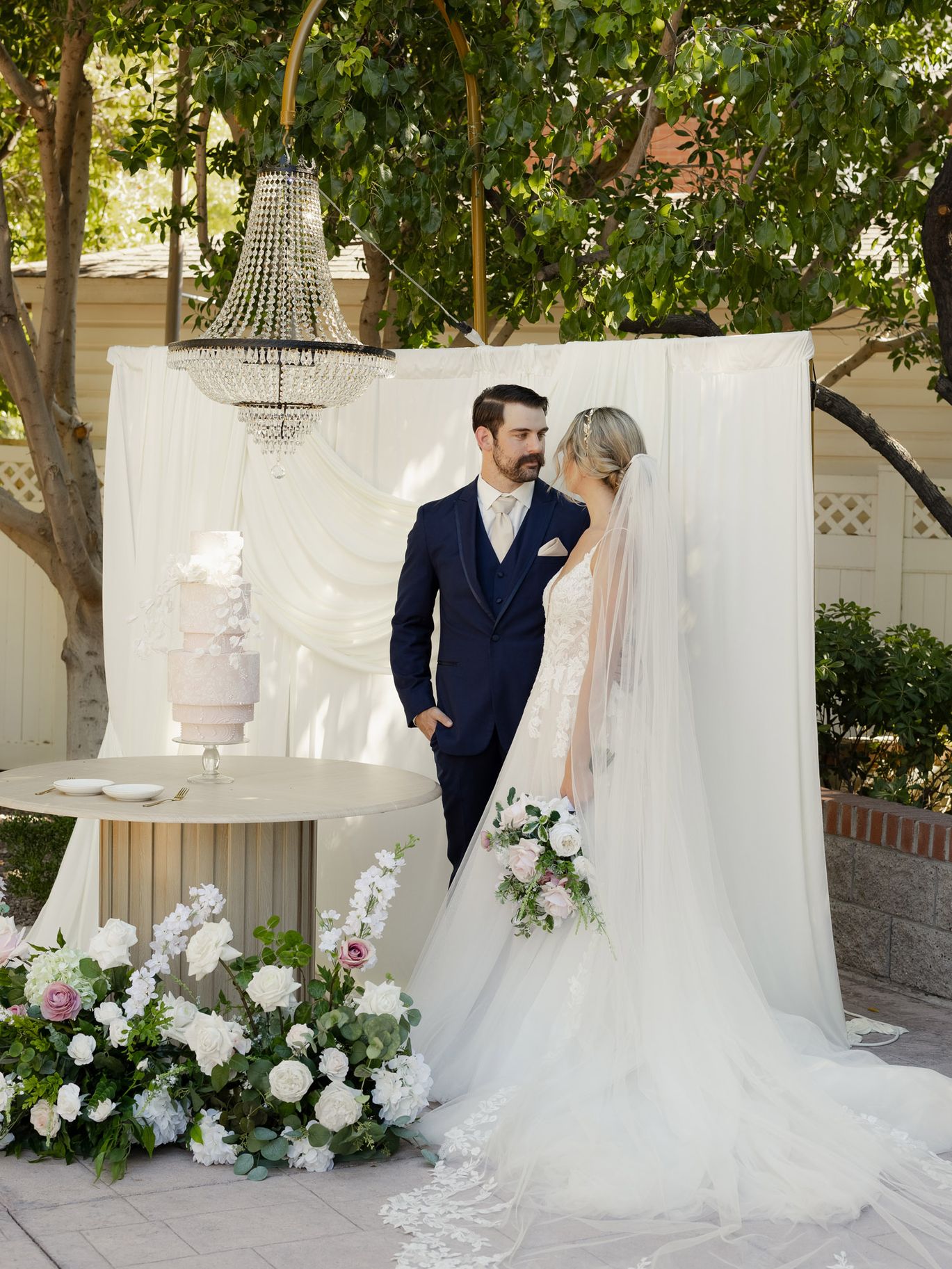 1. Bride and groom posing beside a blush pink wedding cake, crystal chandelier and romantic white and blush floral installation