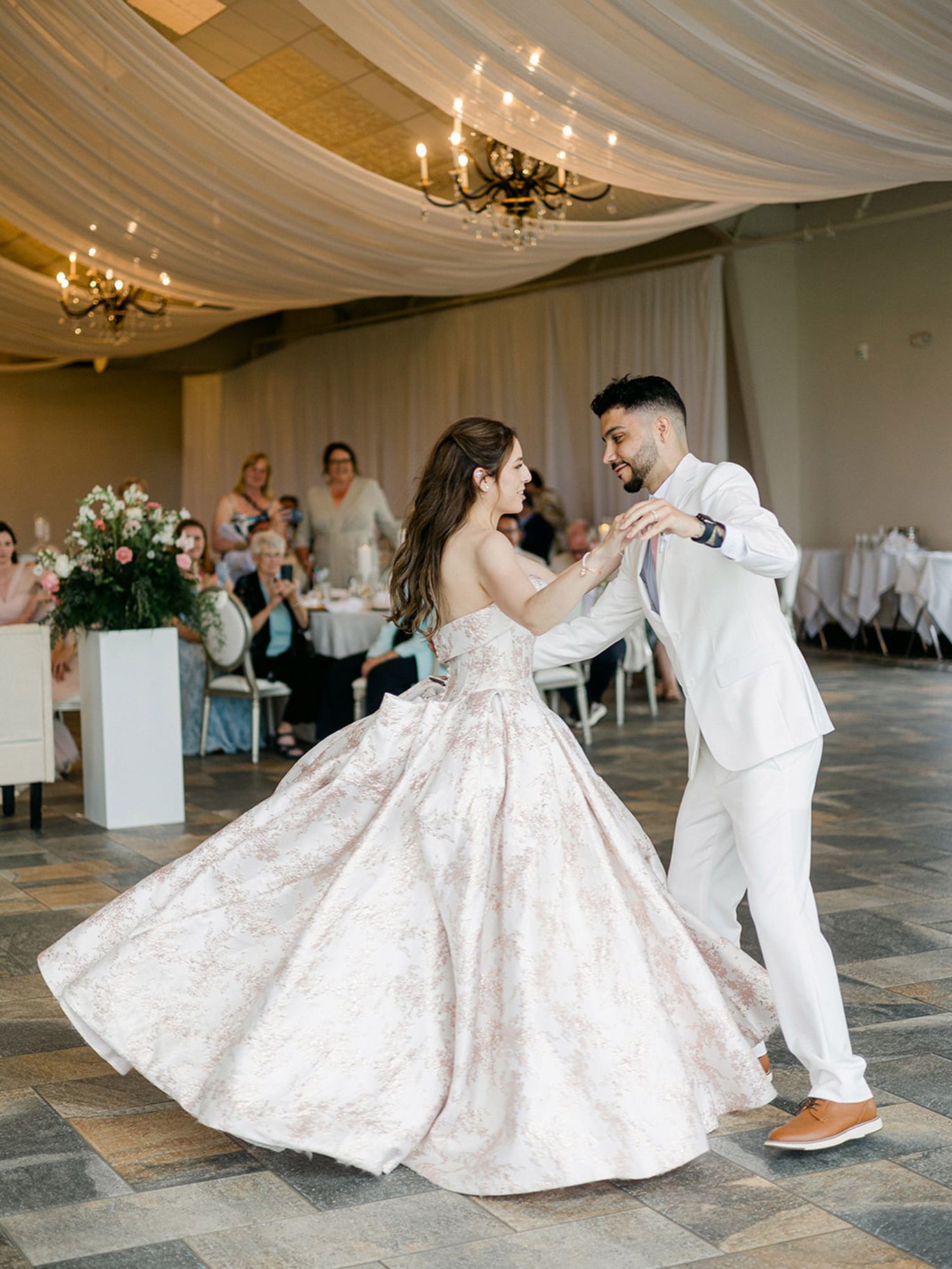 Bride in a full blush-patterned ball gown dances with groom in a white suit beneath draped fabric and chandeliers at their wedding reception.