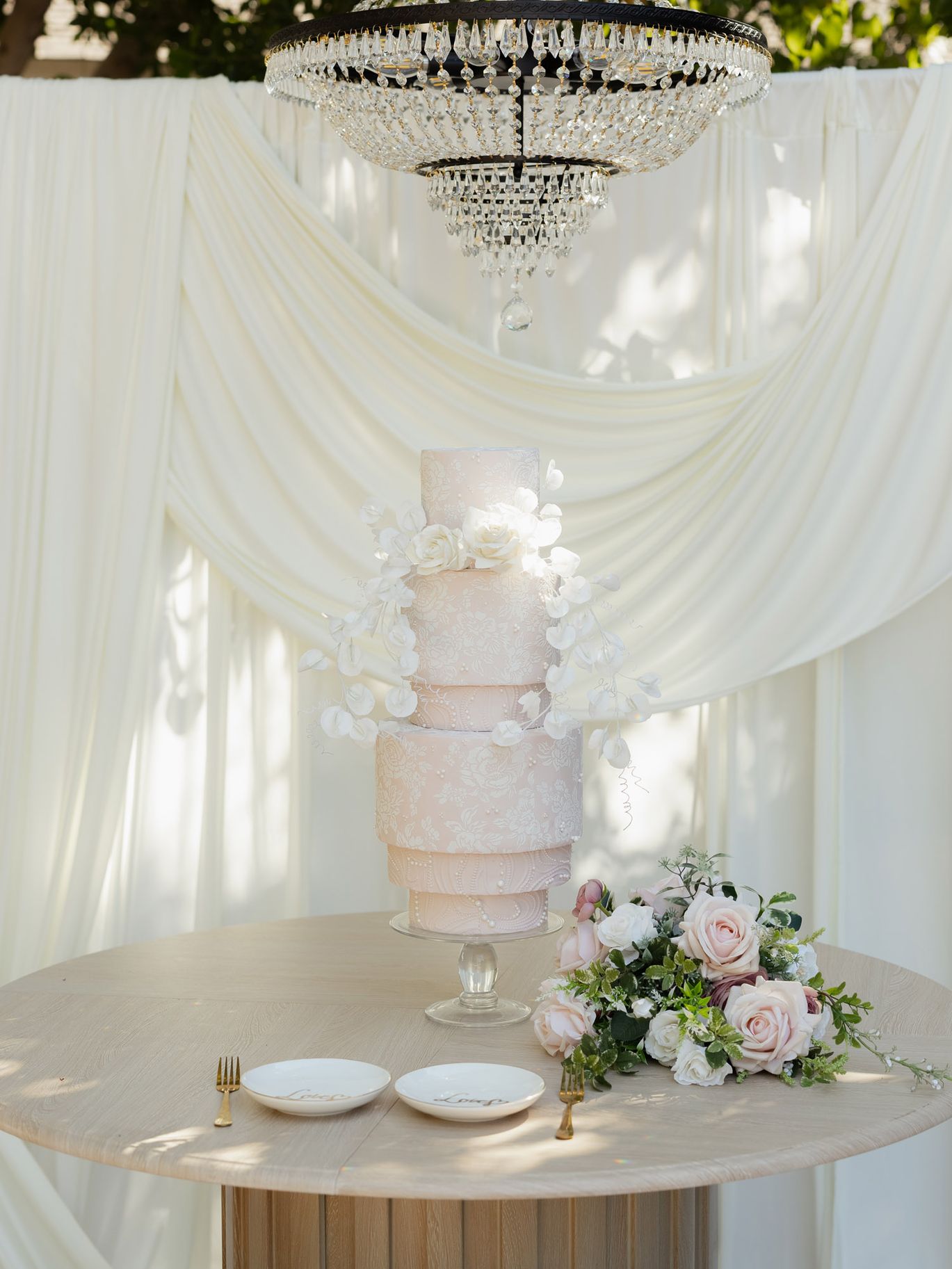 Modern blush pink wedding cake with intricate white floral detailing, cascading flowers and a crystal chandelier overhead