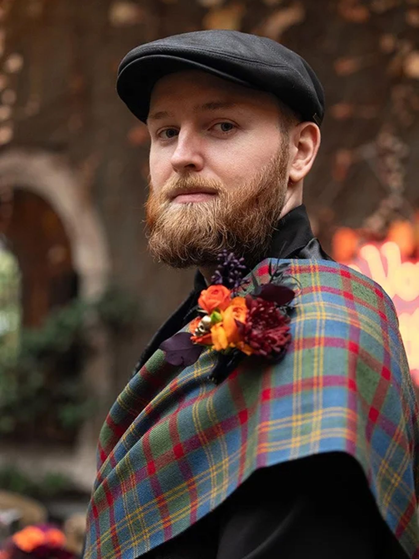 The groom wears a black cap and a colorful tartan sash with a floral boutonniere for the couple’s Halloween wedding.