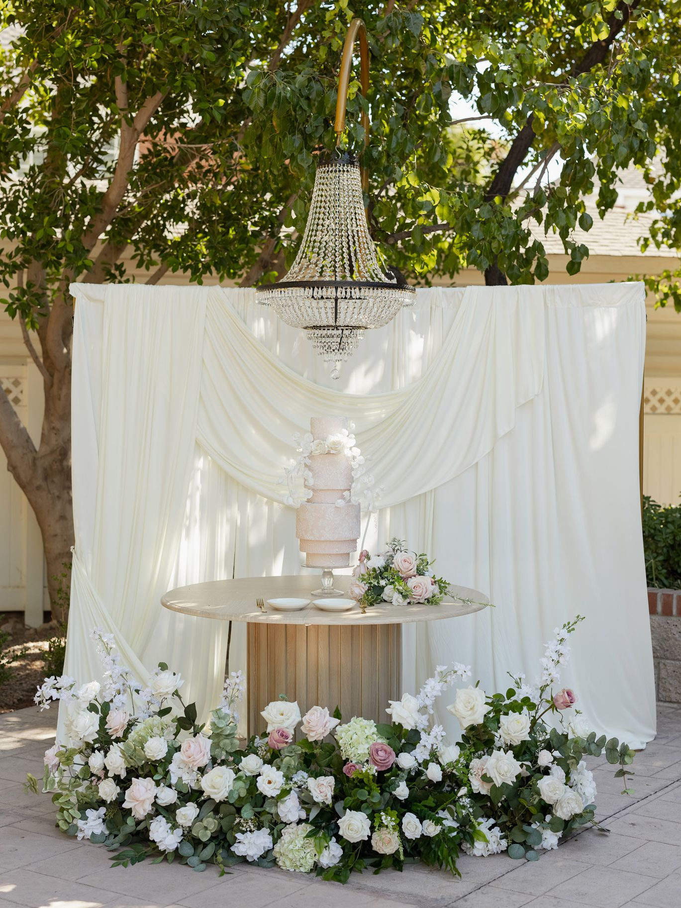 Blush pink wedding cake displayed beneath a crystal chandelier with white draping and romantic floral arrangements