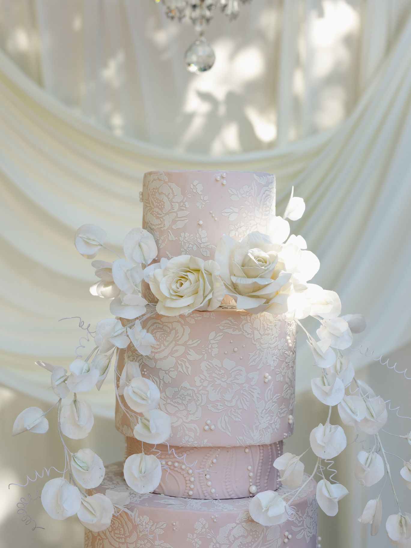Close-up of a blush pink wedding cake featuring intricate white floral patterns, pearl details and cascading white flowers