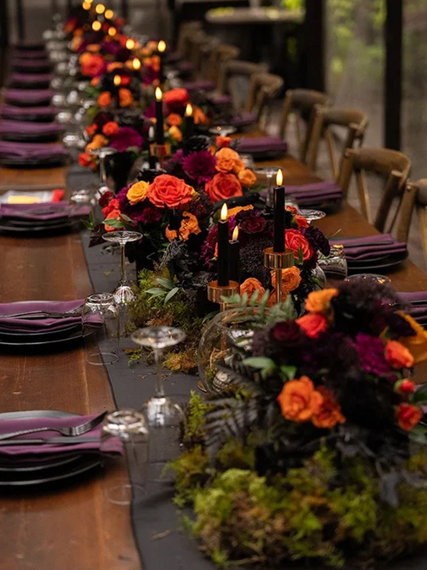 A long reception table at Paige and Dylan’s Halloween wedding features dark place settings, purple napkins, candlelight and colorful floral centerpieces.