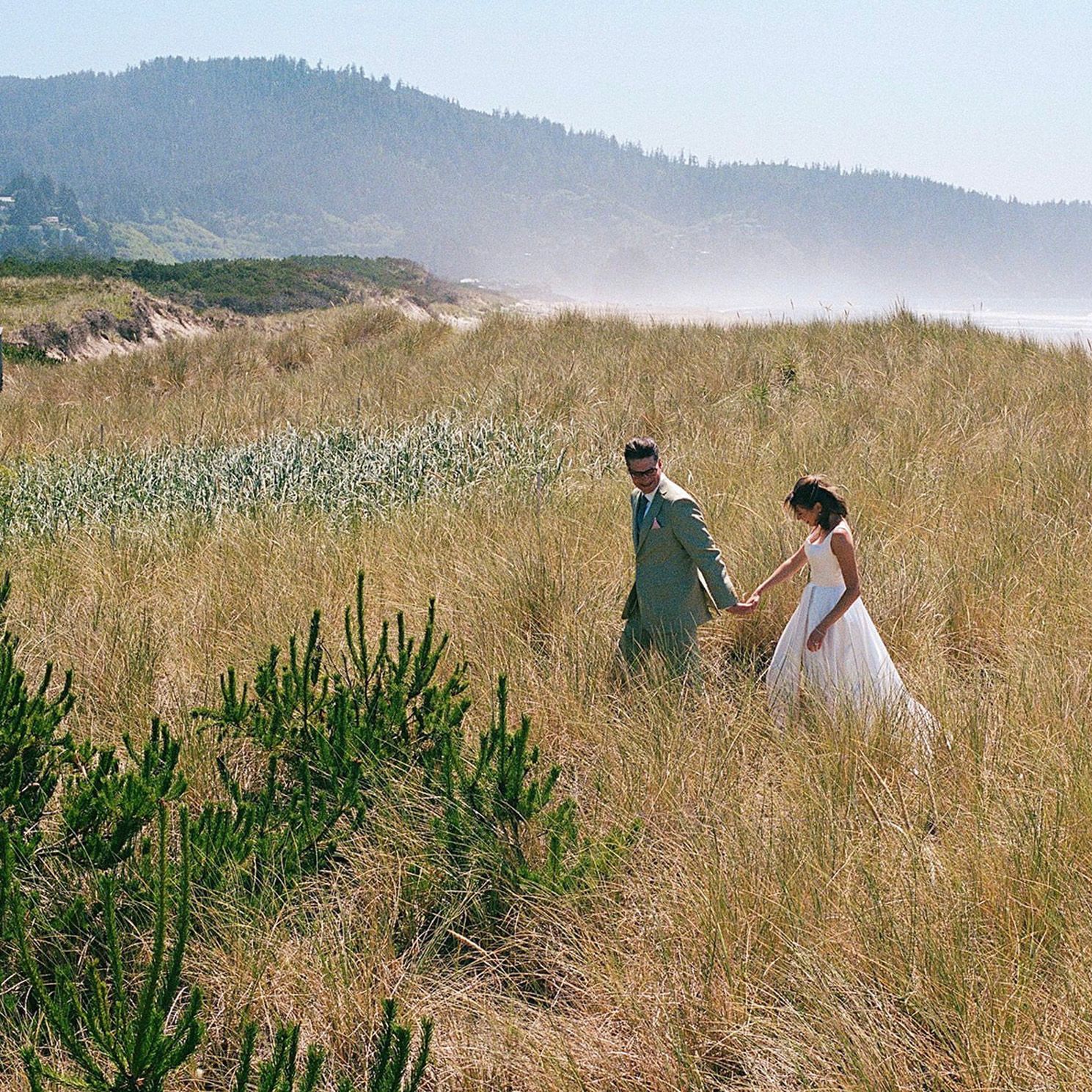Bride and groom walk hand in hand through oceanside field in coastal wedding day