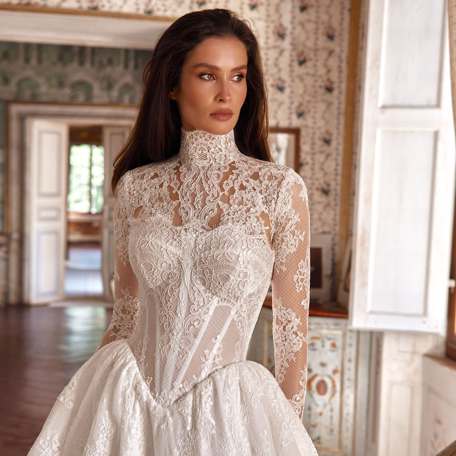Oksana Mukha Emily wedding gown from the Fall 2027 Lumière Heritage collection, featuring a high-neck lace collar, sheer embroidered sleeves and a voluminous floral lace ballgown skirt.