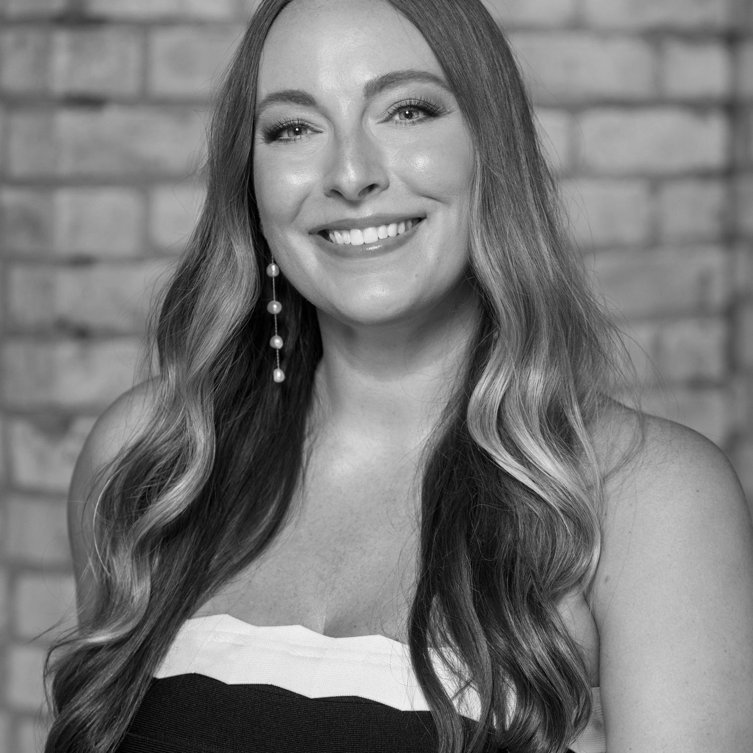 Black and white professional portrait of Jessi Pallan, Creative Director of Iron Diamond Media, wearing a strapless dress with long, wavy hair and pearl earrings, smiling in front of a brick wall.