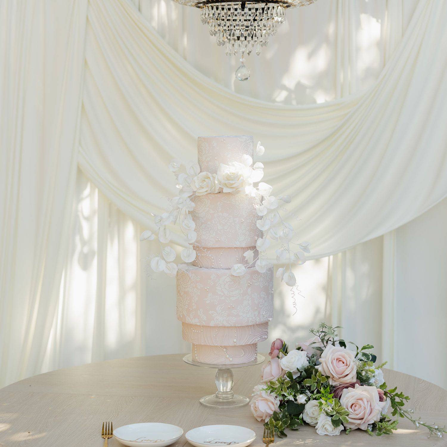 Modern blush pink wedding cake with intricate white floral detailing, cascading flowers and a crystal chandelier overhead