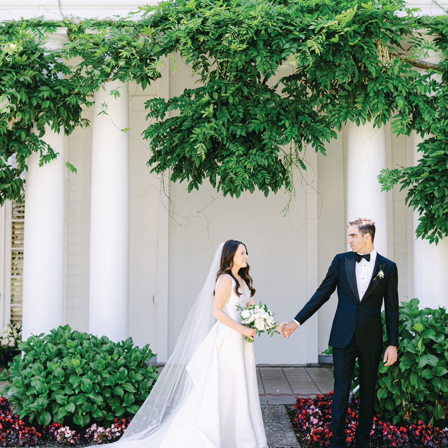Bride and groom hold hands outside white venue with lush greenery