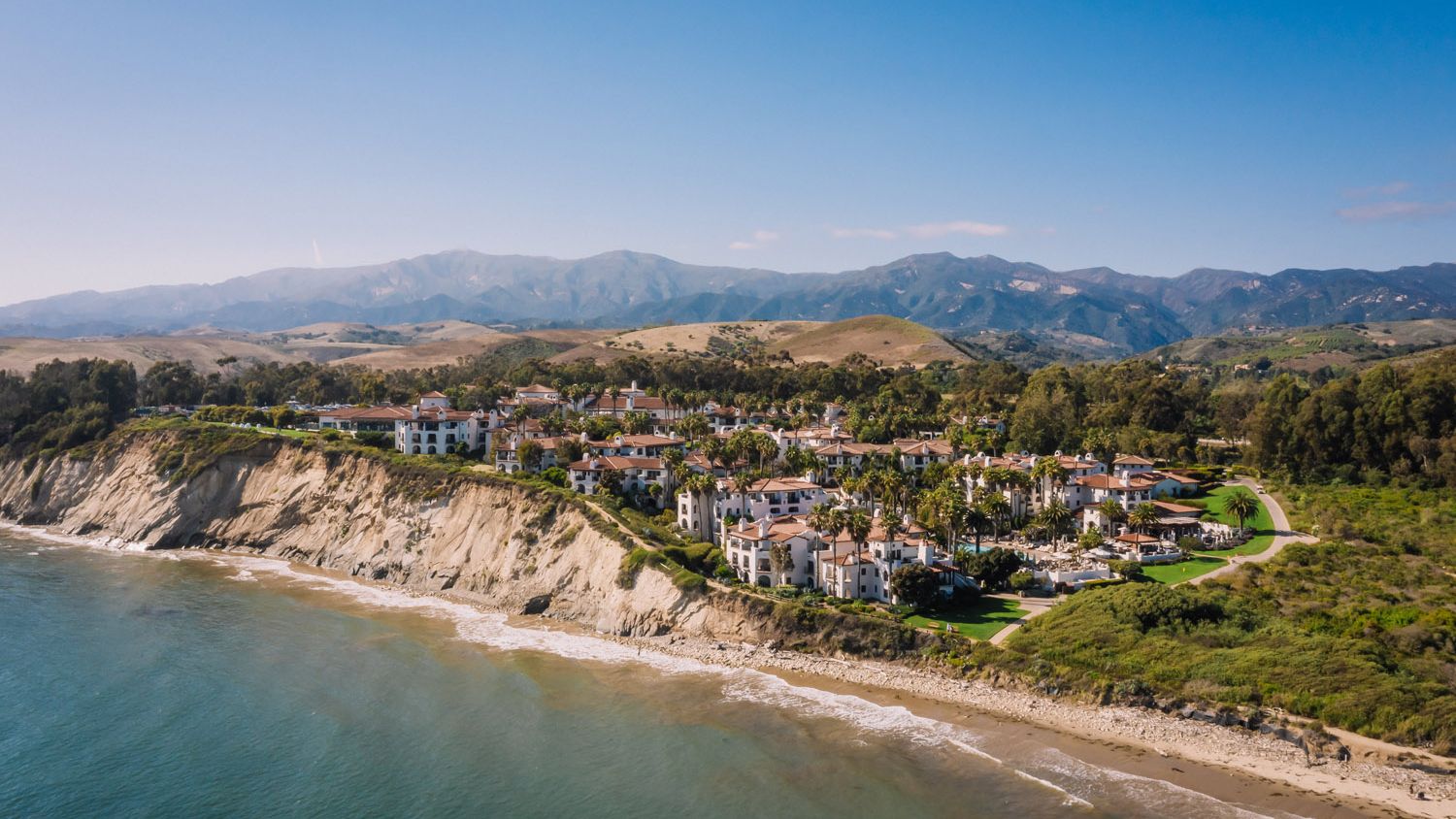 An aerial view of The Ritz-Carlton Bacara, Santa Barbara along the Pacific coastline, with the resort’s Spanish-style buildings set between the ocean and Santa Ynez Mountains