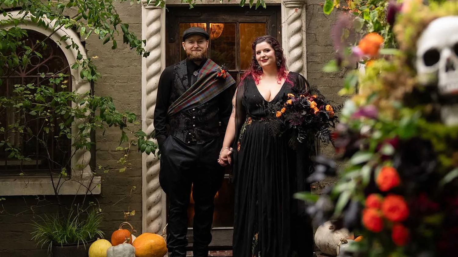 Paige and Dylan pose in black wedding attire outside The Corson Building for their Halloween wedding in Seattle, with pumpkins and fall flowers around the entrance.