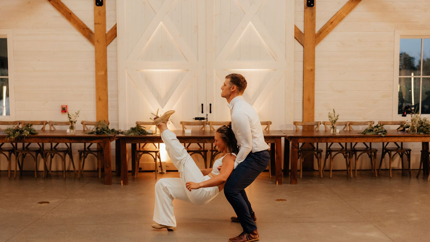 Groom supports his bride in a dramatic seated dance pose, with her legs raised during a choreographed wedding performance in a barn venue.