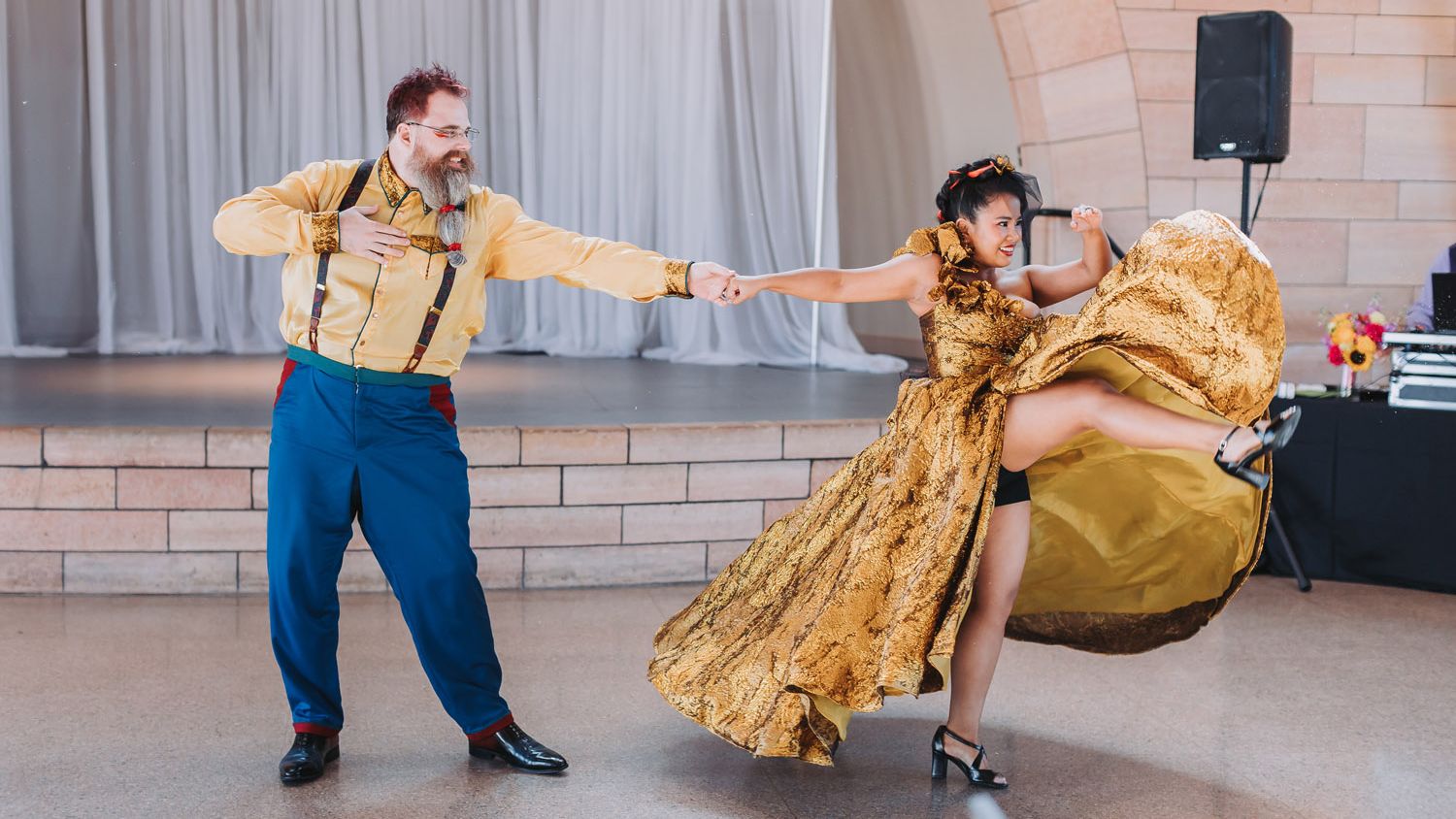 Dance partners perform an energetic routine, with the woman lifting her gold dress into a sweeping flourish as her partner holds her hand.