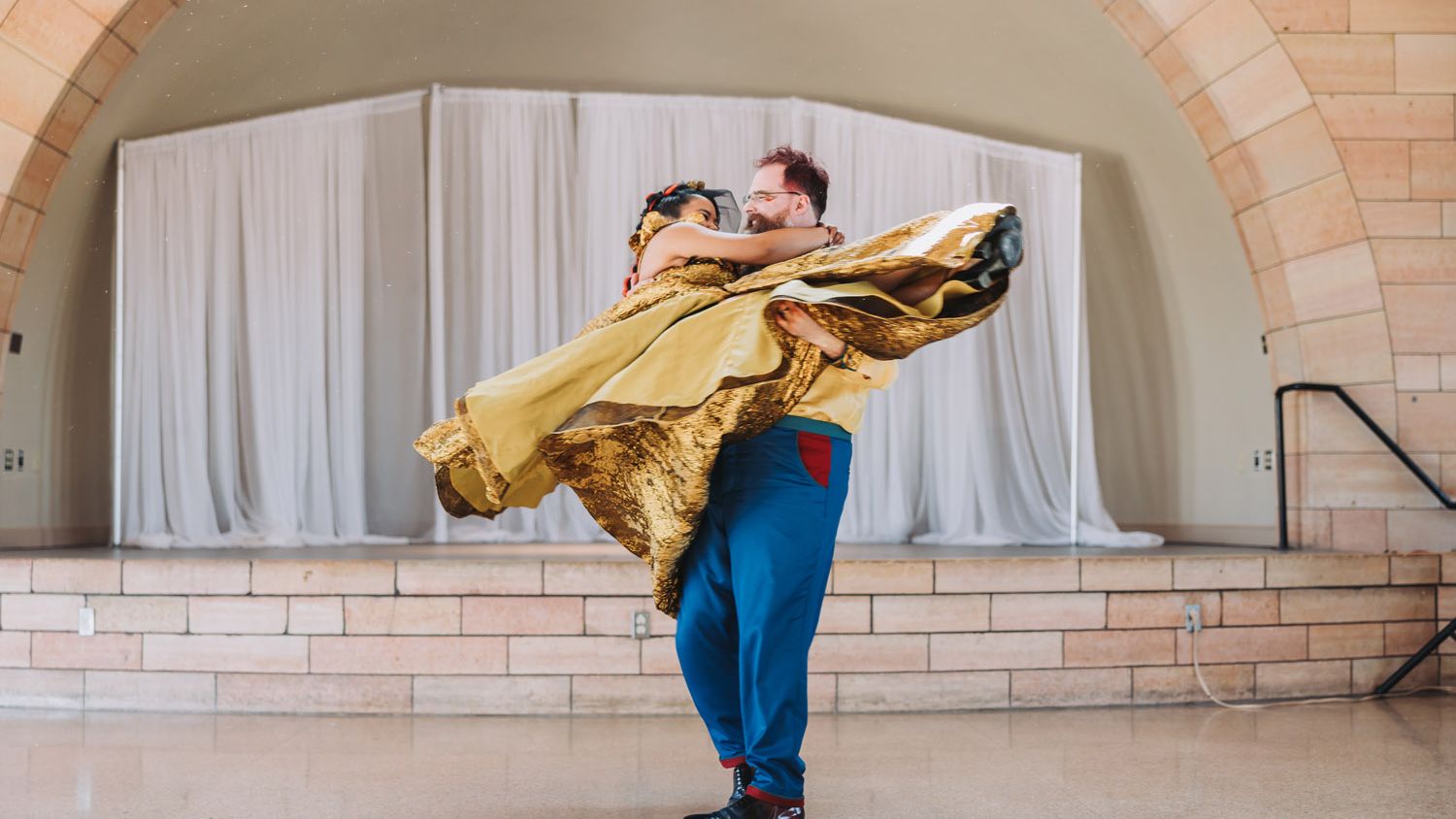 Dance partners perform a dramatic lift, with the woman in a flowing gold dress held aloft in front of a draped stage.