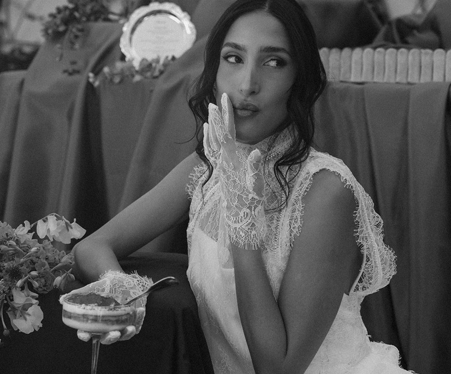 Image of bride with lace gloves on sitting with a desert cup in one hand and the other hand touching the corner of here mouth