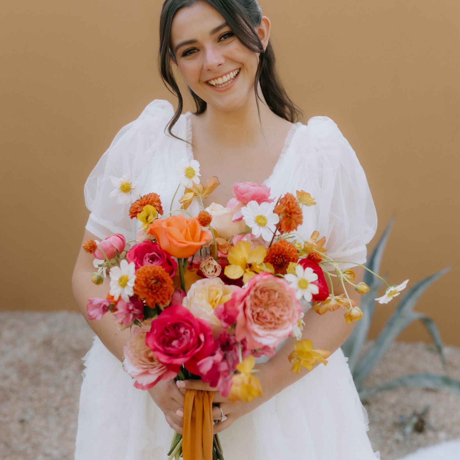 Smiling bride in a white puff-sleeve wedding dress holds a colorful garden-style bouquet of pink, orange and yellow blooms by Fleur de Vie Studio