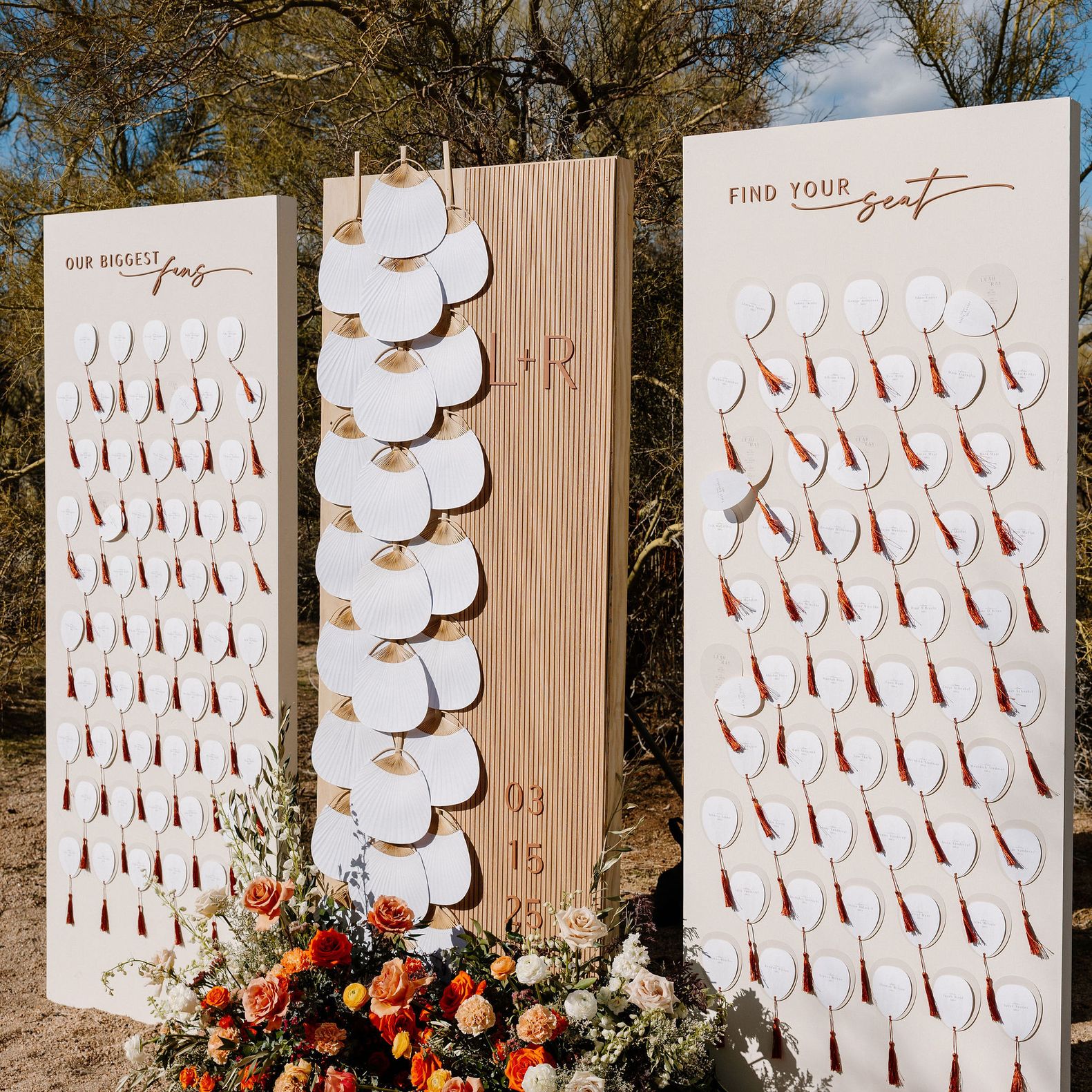 Outdoor wedding escort card display with white fan-shaped place cards, rust tassels and orange, peach and white flowers