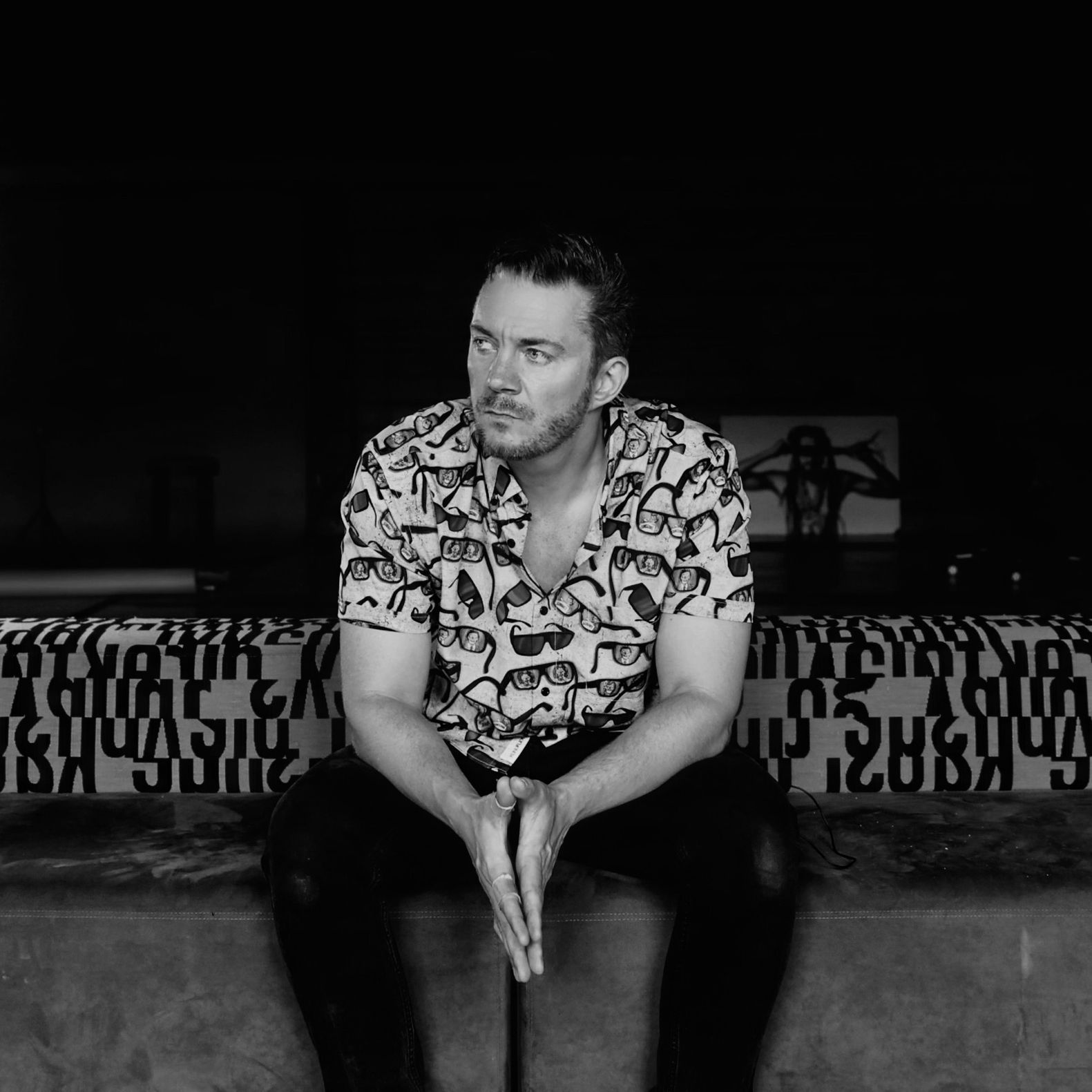 Black-and-white portrait of wedding cinematographer Jonathon Goodro seated in a photography studio, wearing a patterned shirt with lighting equipment visible behind him.