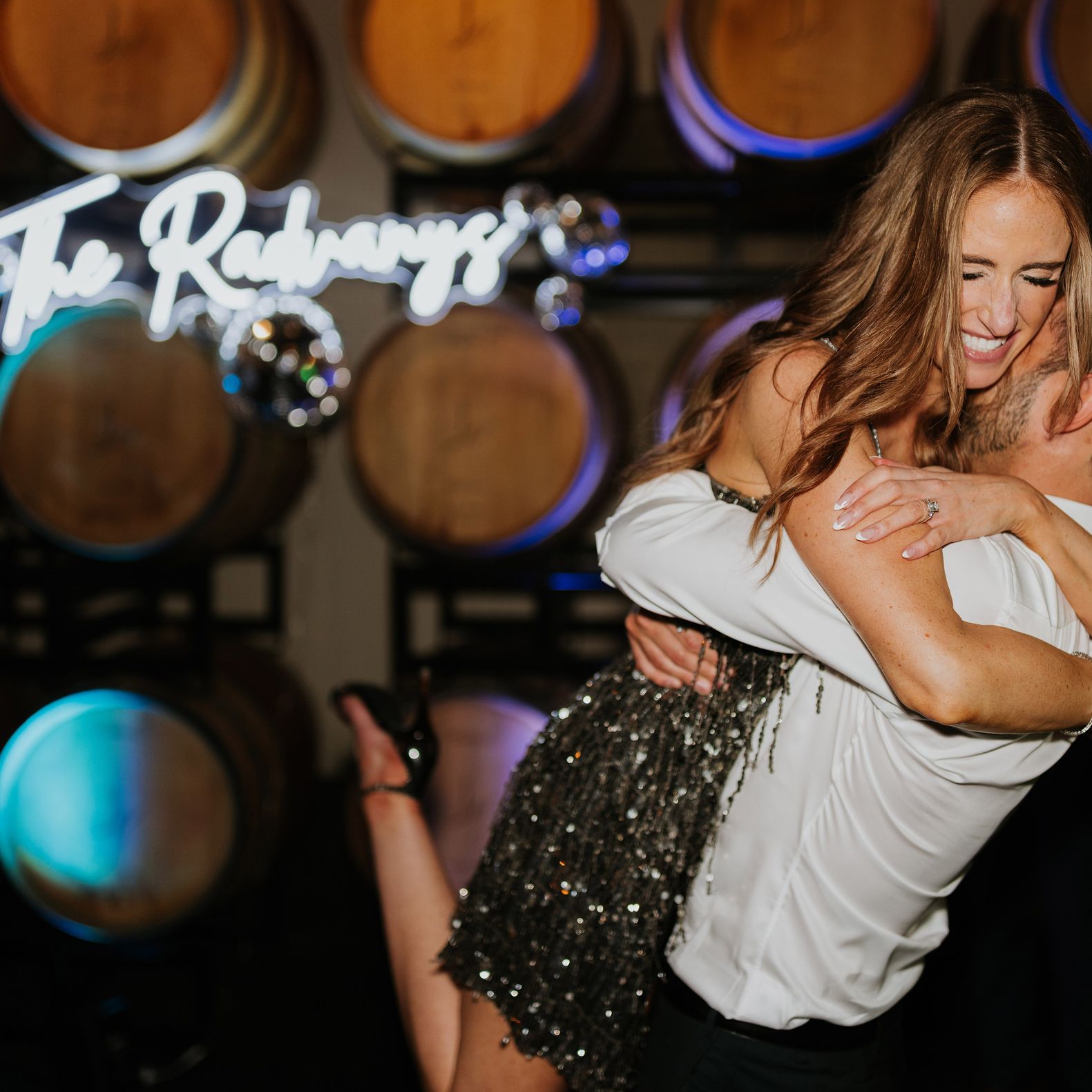 Bride and groom happily embrace in their festive after party outfits in front of a neon sign and wine barrels at JM Cellars