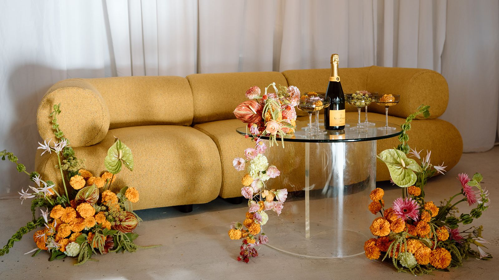 Modern wedding lounge with mustard sofa, mirrored cocktail table, champagne and colorful sculptural floral arrangements
