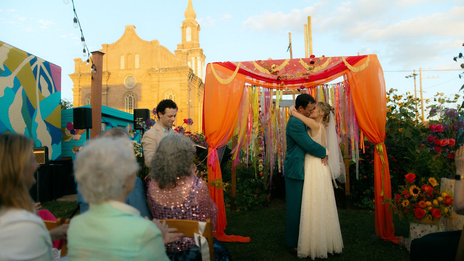 Newlyweds kiss beneath a colorful orange canopy during an outdoor garden ceremony.